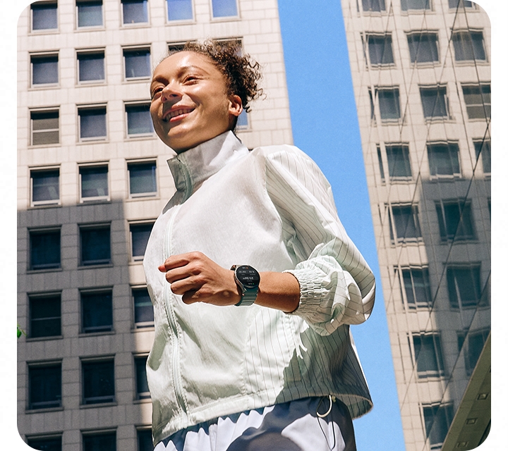 A woman in a workout outfit wearing a Galaxy Watch7 is seen walking on a city block surrounded by buildings.