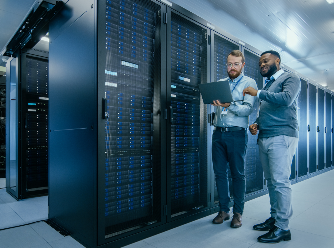 IT Technician in Glasses with a Laptop Computer and Engineer Colleague are Talking in Data Center while Working Next to Server Racks