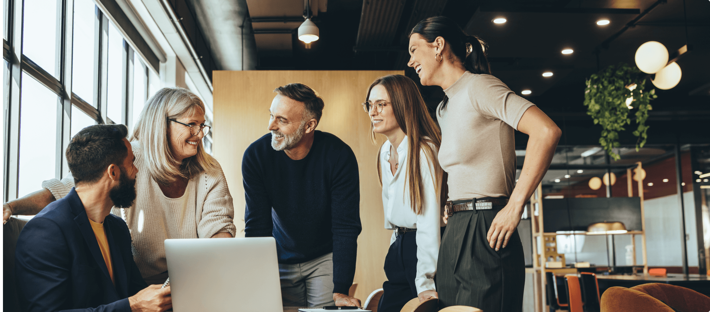 Colleagues standing around a table smiling and looking at each other in front of a laptop computer