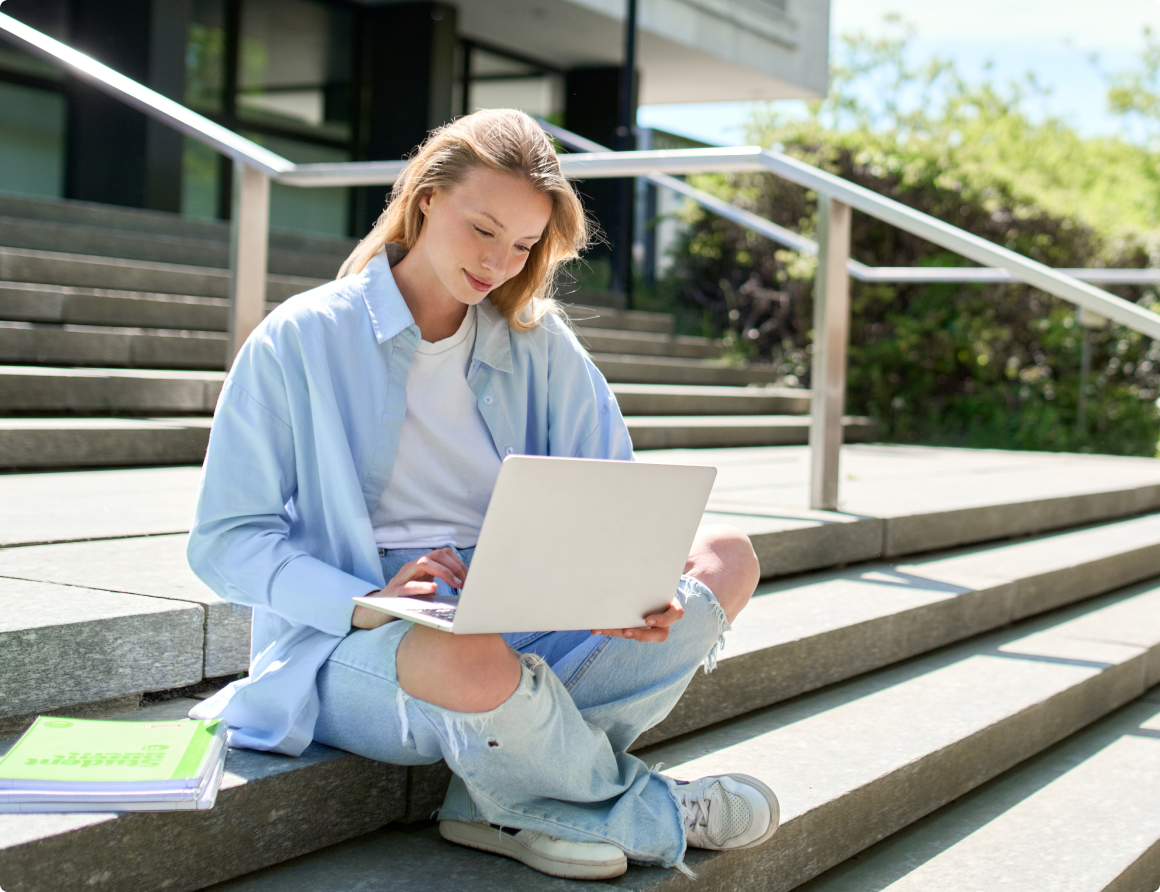 Photo of a student sitting on the stairs of a building working on her laptop