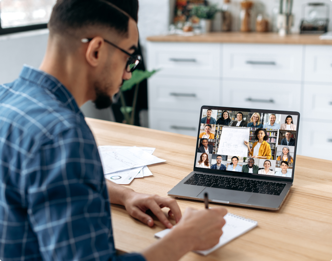 Man is working on his laptop and taking notes on his notepad while he is in a group video call