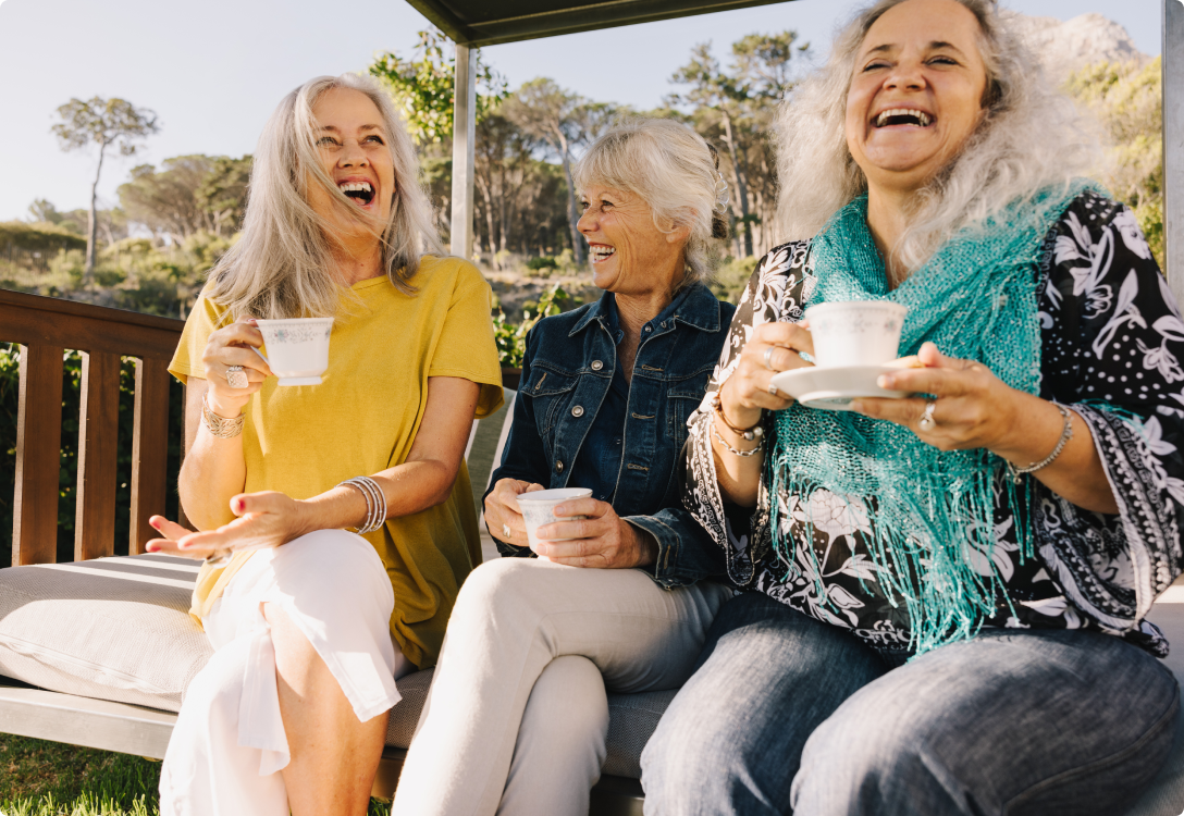 Photo of three ladies laughing and drinking tea outside on a bench