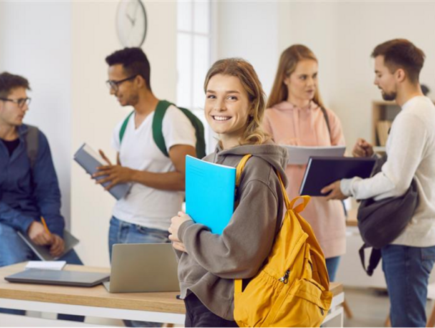 Photo of a college student holding a blue book and wearing a yellow backpack in class